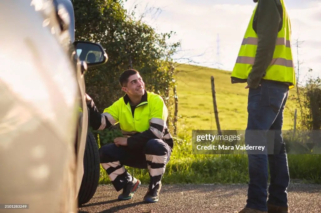 Recovery technician in high-visibility clothing crouching beside a broken-down vehicle at the roadside.